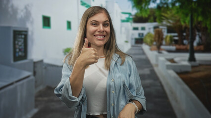 Woman wearing blue denim shirt and white crop top waves hands on street amid sunlit sidewalk by white buildings; joy.