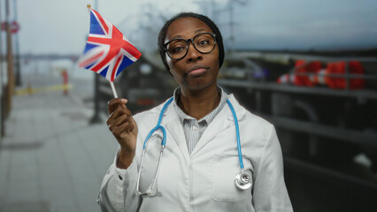 Woman doctor outdoors holding a british flag in an urban street setting, wearing glasses and a...