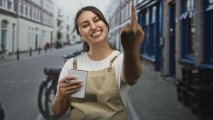 Woman waitress in beige apron holds notepad and pen and shows middle finger while smiling on city street; defiance.