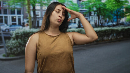 Fototapeta premium Woman hand to forehead on street in front of row houses and hedges, lips pursed gesture wearing brown sleeveless top; annoyance.