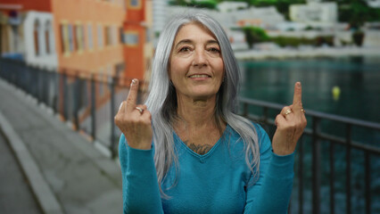 Woman with grey hair makes expressive gestures on seaside promenade, wearing blue sweater, surrounded by beach scenery, embodies outdoor freedom and defiance in vibrant setting.