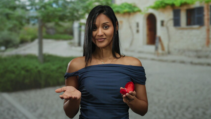 Woman gesturing shrug outdoors with ring box in hand on cobblestone street with expression of uncertainty in casual dress in a village setting.