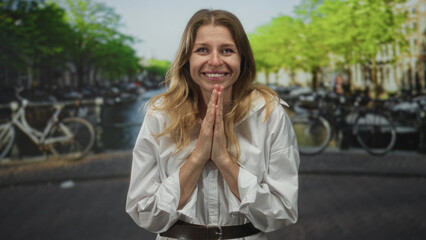Woman with hands pressed together, smiling on a street by an amsterdam canal and water in the city outdoors; quiet gratitude.