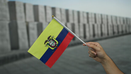 Hand holding ecuadorian flag outdoors near gray monument wall in urban area, symbolizing pride and...