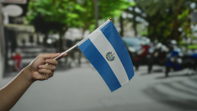 Hand holds el salvador flag outdoors on a sunny day in a lively city street, representing national pride, culture, and identity.