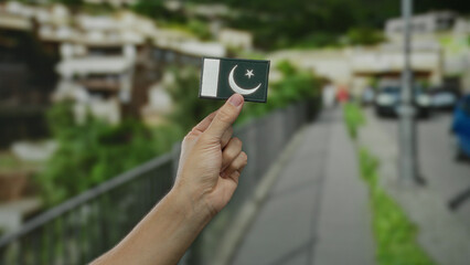 Caucasian man holding an embroidered pakistan flag patch on an urban street, showcasing national pride in an outdoor city setting.