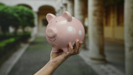 Hand holds pink piggy bank in an old university courtyard highlighting financial themes amidst...