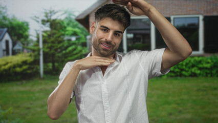 Young hispanic man smiling makes framing gesture with hands at building entrance; confidence and joy.