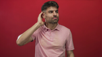 Man scratches his head and tilts his shoulder against a bright red studio wall, offering a puzzled...