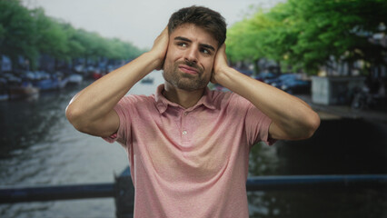 Man covers ears with both hands on canal street bridge overlooking moored boats and green trees;...