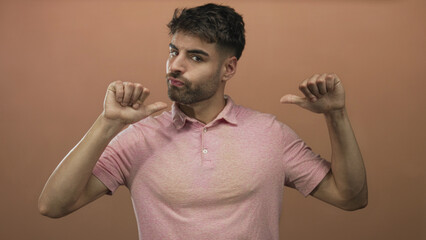 Hispanic man wearing pink shirt pointing both thumbs at chest and pouting lips against peach studio wall; confidence.