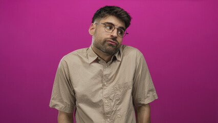 Obraz premium Young hispanic man shrugs shoulders with raised brow and tilted head in bright pink studio; uncertainty.
