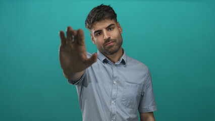 Young hispanic man pushes palm outward at camera in teal studio setting with serious expression; defiance.