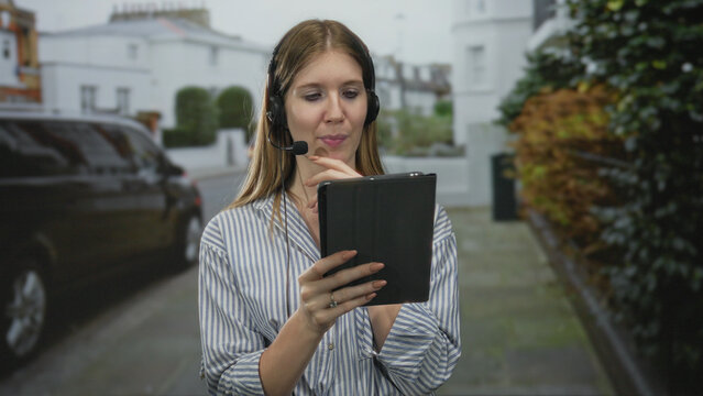 Young woman wearing headset holds tablet with finger pointing at screen on street sidewalk; confusion.