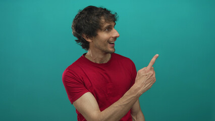 Young man with outstretched hand pointing finger to the right in a studio with teal background; surprise curiosity.