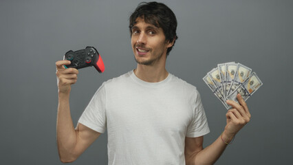 Smiling young man grips black and red game controller and fans us dollar bills in grey studio; success.