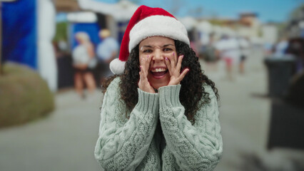 Woman yelling and smiling at street market wearing santa hat and green sweater with people in...