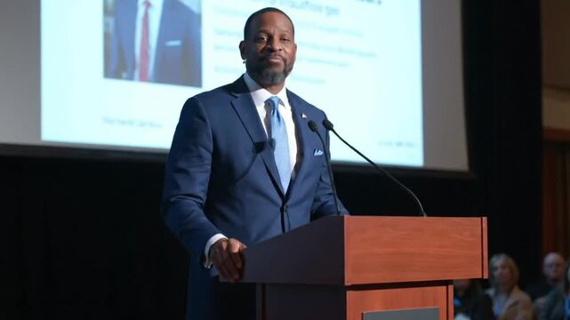 Man in suit presents at podium with mics screen behind