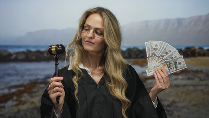 Blonde woman judge in uniform holding us dollars and gavel at a seaside beach setting, capturing a...