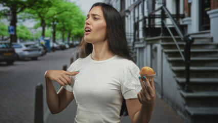 Woman holding muffin and pointing finger on urban street sidewalk amid parked cars and brick townhouses; confusion.