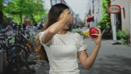 Hispanic young woman covering her eyes with hand holds red apple on bustling urban street;...