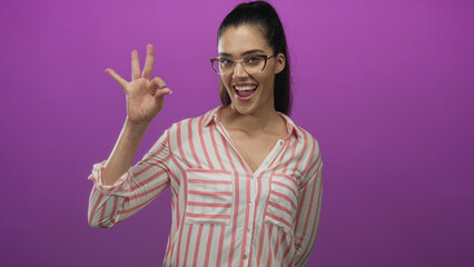 Hispanic woman in striped shirt making ok sign gesture in purple studio with a bright smile; approval optimism happiness confidence.