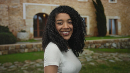 Woman displays curly hair, smiles warmly and wears a white t shirt at building entrance outdoors;...