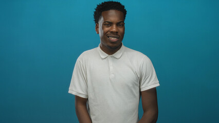 Young african american man wearing white polo squints eyes in blue studio with subtle head tilt;...