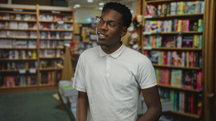 African american man wearing white polo shirt tilts head toward bookshelf in building; intellectual curiosity.