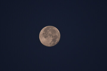 A full moon against a deep blue night sky, with a slightly hazy atmosphere. The moons details are visible, giving it a soft, warm glow.