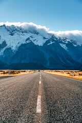 Straight asphalt road leading to snow capped mountain under blue sky among golden field in New Zealand
