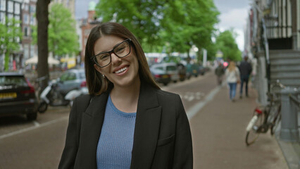 Fototapeta premium Woman smiling on amsterdam canal street, brunette adult in glasses enjoys a sunny day featuring urban architecture and parked cars along historic canals in the netherlands.