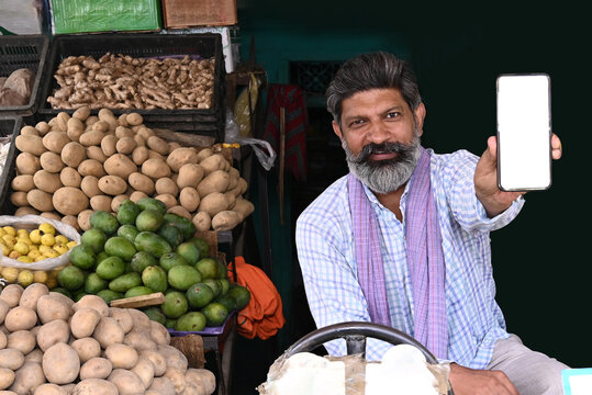 Indian veggie vendor showing empty white screen smartphone for advertising online grocery promotions and easy online grocery payment process at his store on the streets of a market in the city