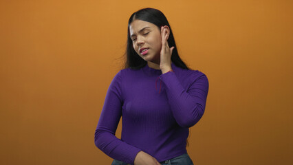 Woman pressing ear with hand against orange wall in studio setting with pained expression;...