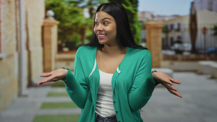 Woman in green cardigan with raised palms shrug pose in front of a modern building facade; uncertainty.