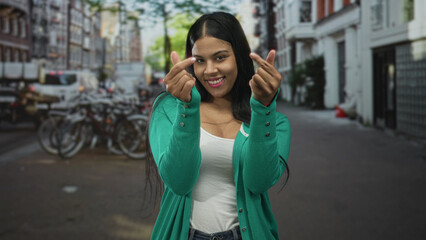 Fototapeta premium Young latin woman makes finger heart gestures with both hands on busy city street; joy connection warmth.