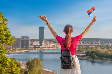 Young woman celebrating with arms raised and waving Serbian flag in Belgrade, overlooking cityscape and Sava river