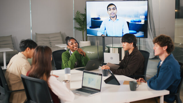 Diverse office teamwork meeting in modern startup with young professionals collaborating around table and video conference screen showing remote colleague