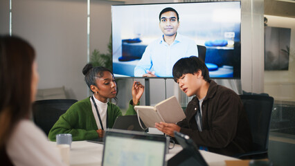 Diverse office teamwork in startup environment with colleagues collaborating in meeting room while remote worker joins via video call creating focused and productive atmosphere