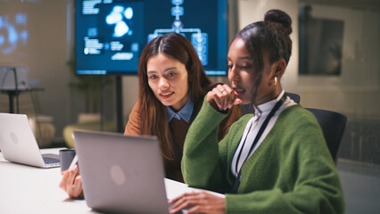 Diverse office teamwork in startup environment as two women collaborate on project using laptop with technology and innovation in modern workspace