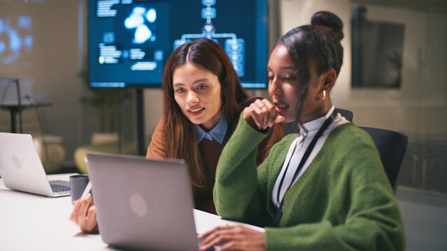 Diverse office teamwork in startup environment as two women collaborate on project using laptop with technology and innovation in modern workspace - Powered by Adobe