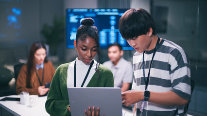 Diverse office teamwork in startup environment with young professionals collaborating on project using laptop modern workspace and digital screen in background