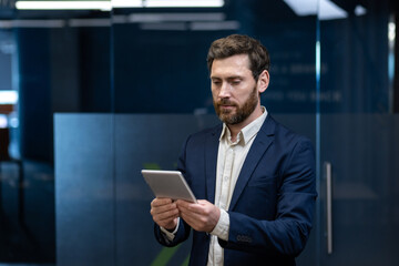A young successful businessman in a suit is standing in an office building and using a tablet he is holding in his hands