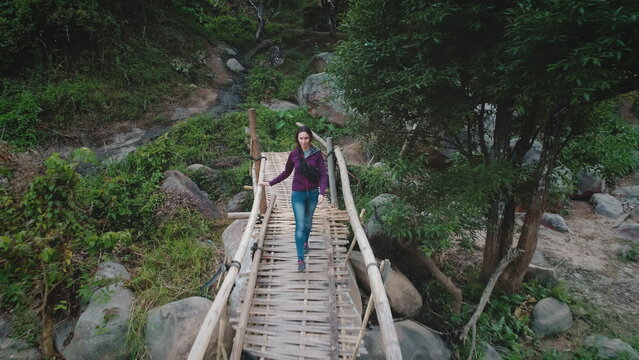 Aerial drone footage of a female tourist walking on a bamboo bridge across a small stream in a tropical forest, enjoying the tranquility of nature