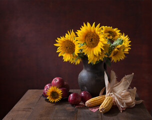 Rustic autumn still life.  Apples, Corn cobs  and bouquet of Sunflowers in clay pot on old rural wooden table.