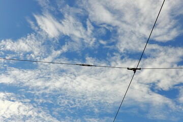 Blue sky with white clouds intersected by tram or trolleybus wires, with city buildings below. Urban infrastructure and abstract cityscape concept.