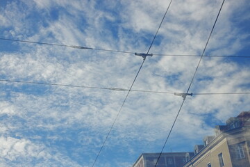 Blue sky with white clouds intersected by tram or trolleybus wires, with city buildings below. Urban infrastructure and abstract cityscape concept.