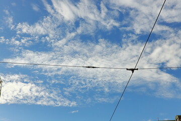 Blue sky with white clouds intersected by tram or trolleybus wires, with city buildings below. Urban infrastructure and abstract cityscape concept.
