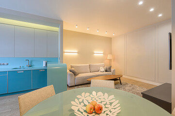 modern living room features a blue kitchen area, beige sofa, glass table with fruit. Warm lighting illuminates the room