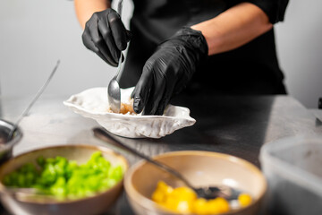 Chef in black gloves preparing food in a professional kitchen, mixing ingredients in a white bowl with chopped bell peppers nearby. Hygienic culinary process on stainless steel surface.